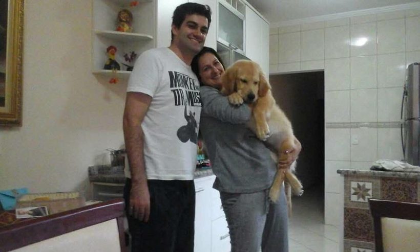 Anderson e Verônica na cozinha de casa; ela segura o cachorro no colo.