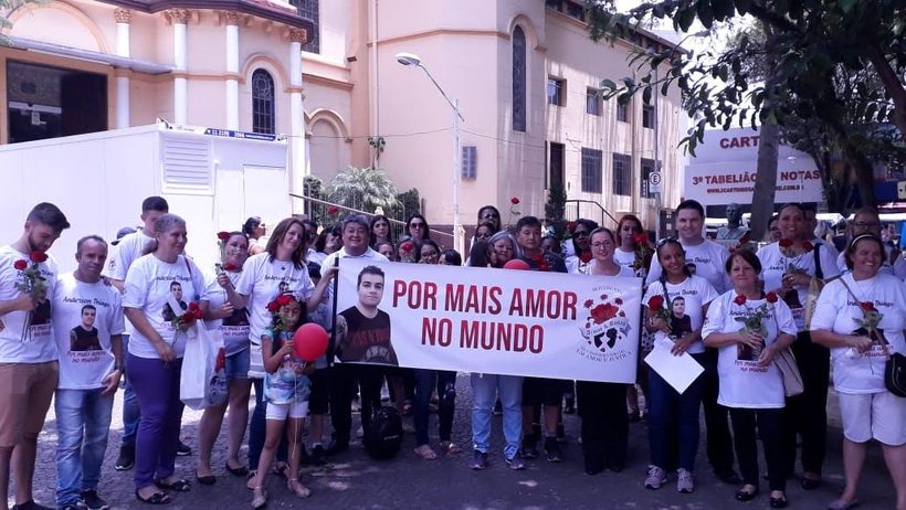 Familiares e amigos vestidos de branco, com rosas vermelhas, seguram a faixa “Por mais amor no mundo”.