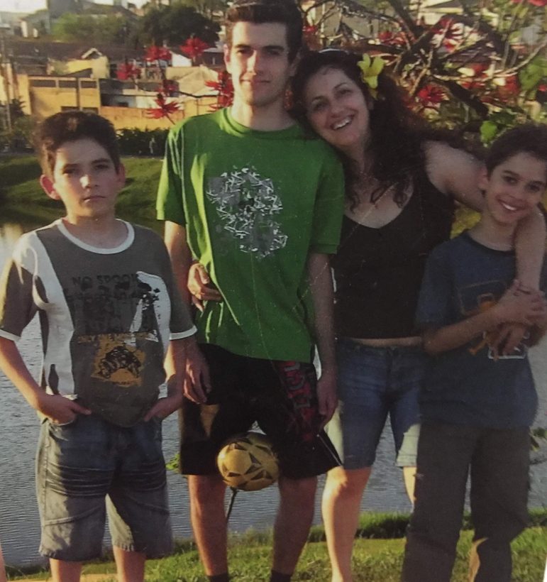 Foto antiga: Verônica abraçada aos três filhos à beira de um lago. Anderson, o mais alto, está de camiseta verde.